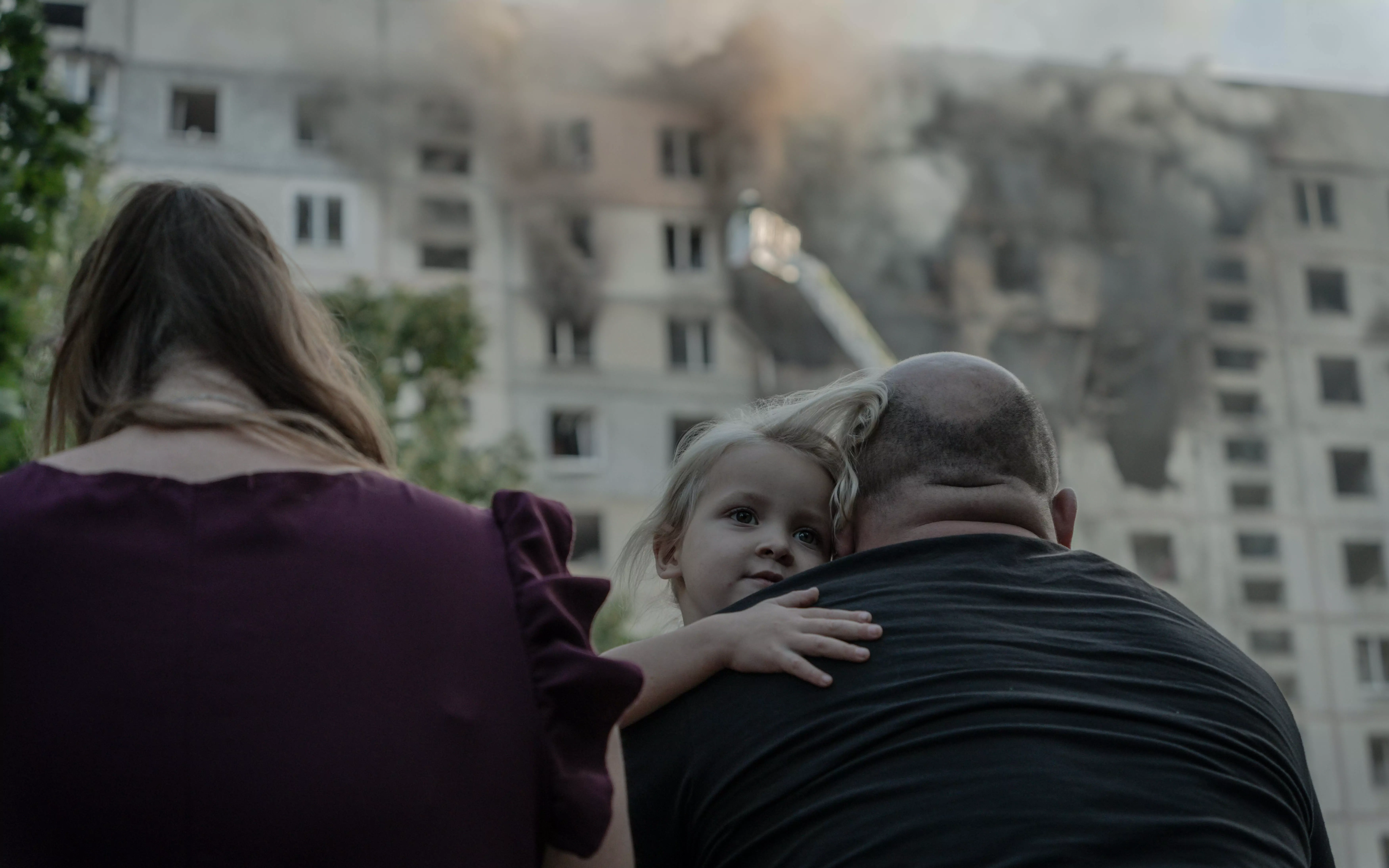 Дівчинка в обіймах батьків на фоні палаючої багатоповерхівки у Немишлянському районі після влучання ворожого КАБу, 15 вересня 2024 / Фото: Іван Самойлов, Ґвара Медіа