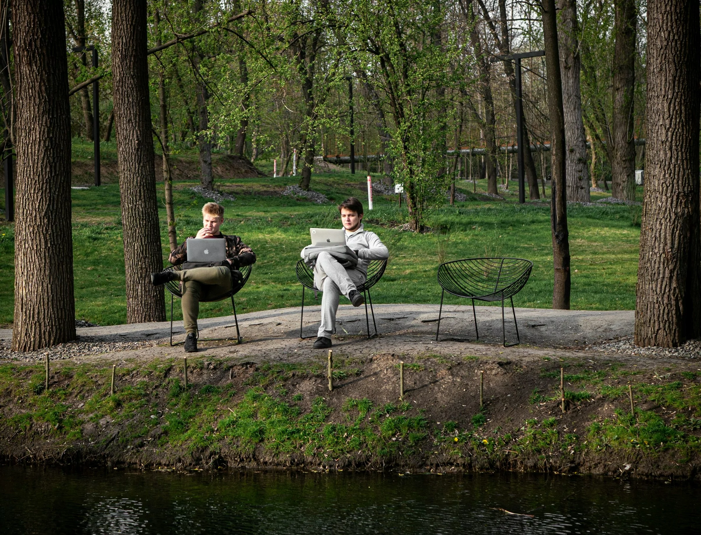 Two people working remotely on laptops while sitting near a quiet park creek.