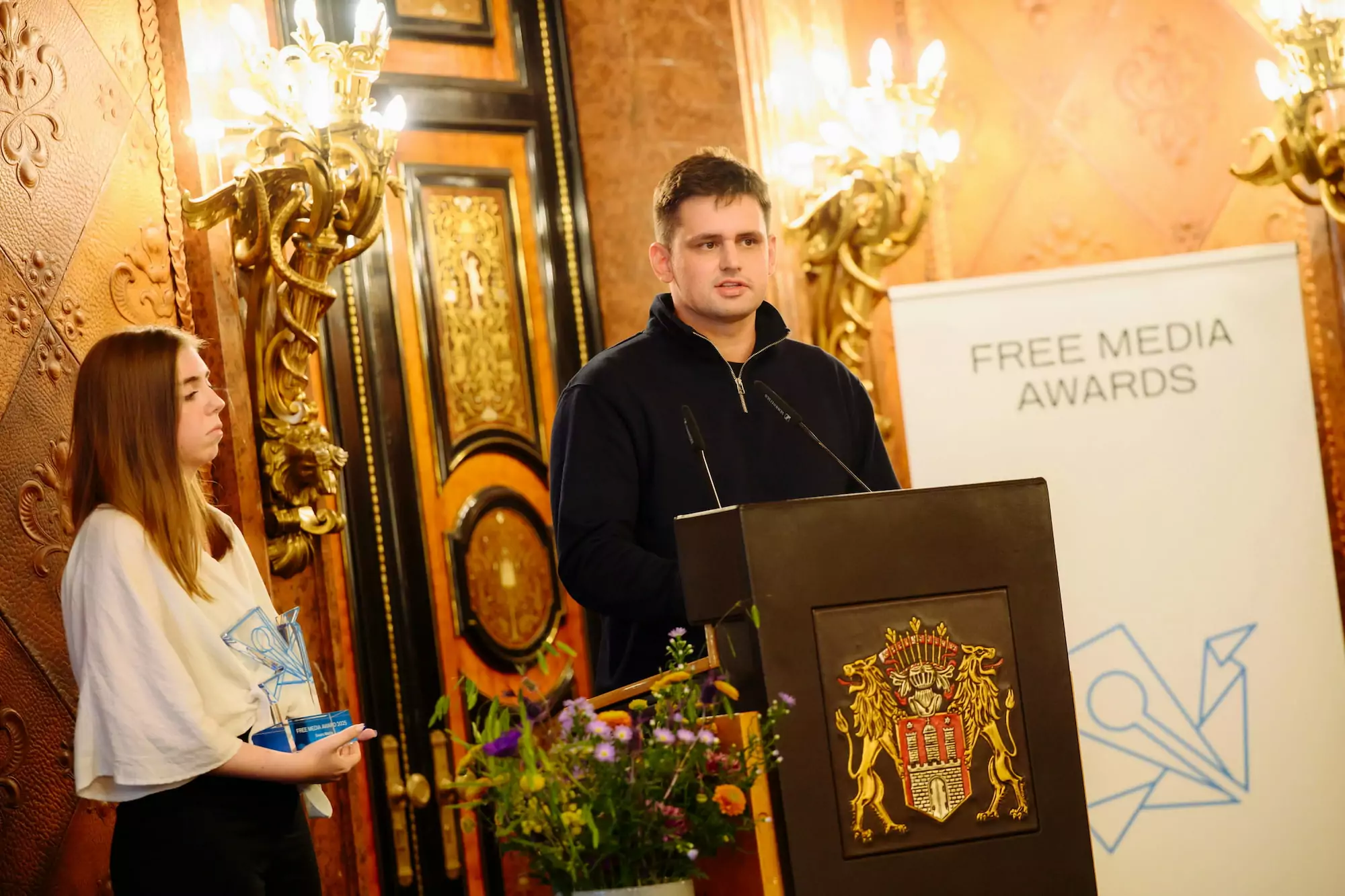 Woman in white blouse (on the left) and man in the black jumper (on the right) who's giving a speech