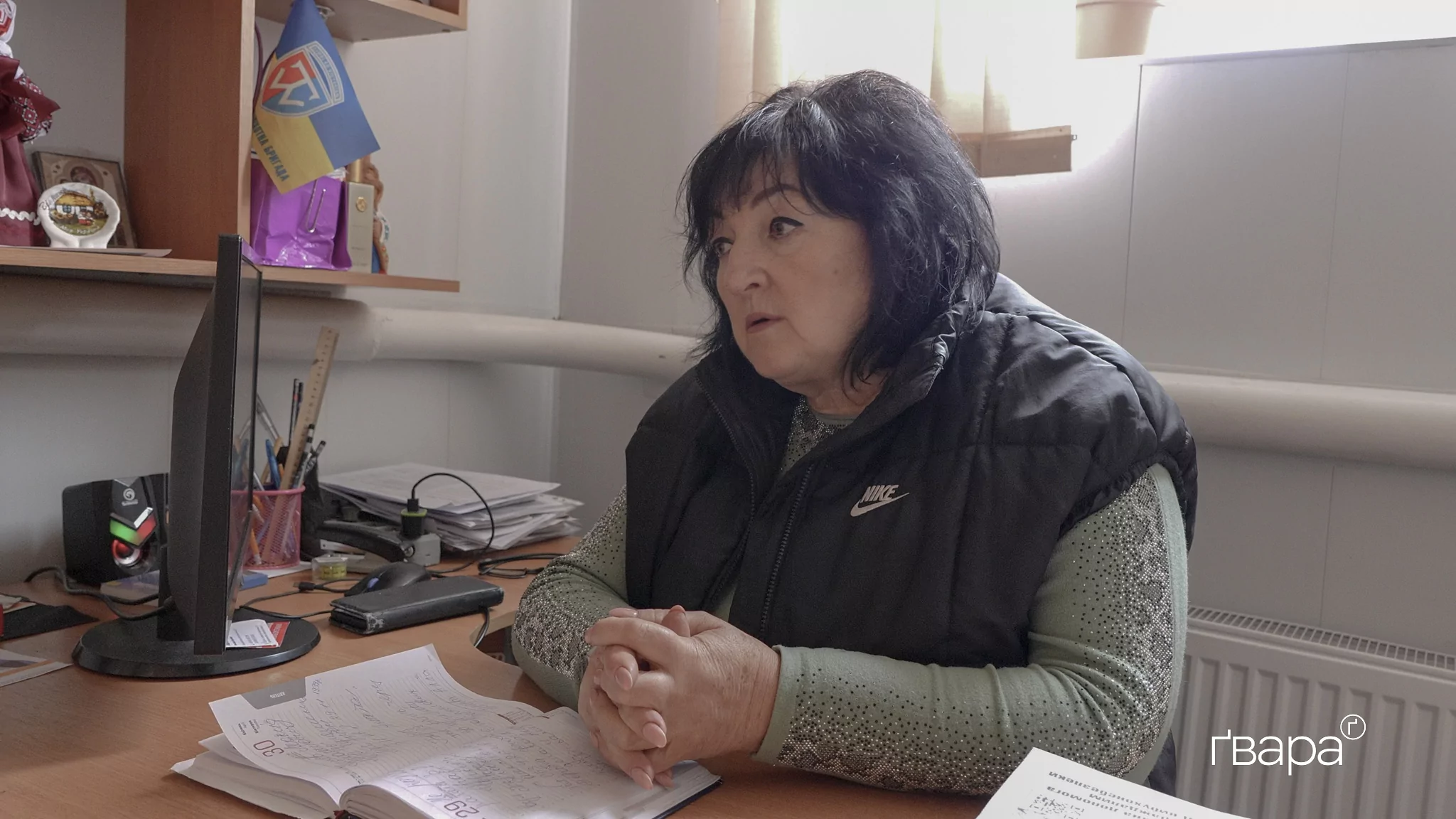 Жінка в зеленій кофті і чорній жилетці сидить за столом у світлій кімнаті