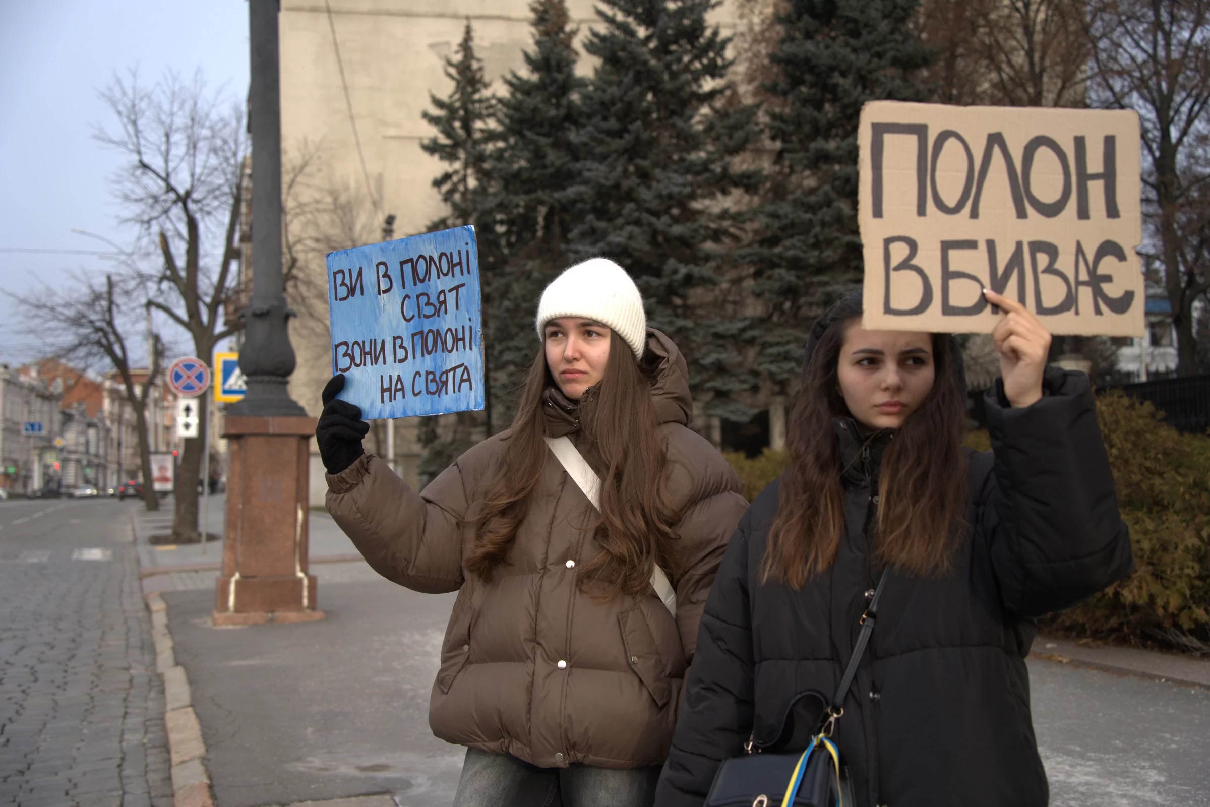 Two women standing on the sidewalk and holding signs for a demostration in Kharkiv. "You're captivated by holidays; they're in captivity on holidays" (on the left), "Captivity kills" (on the right).