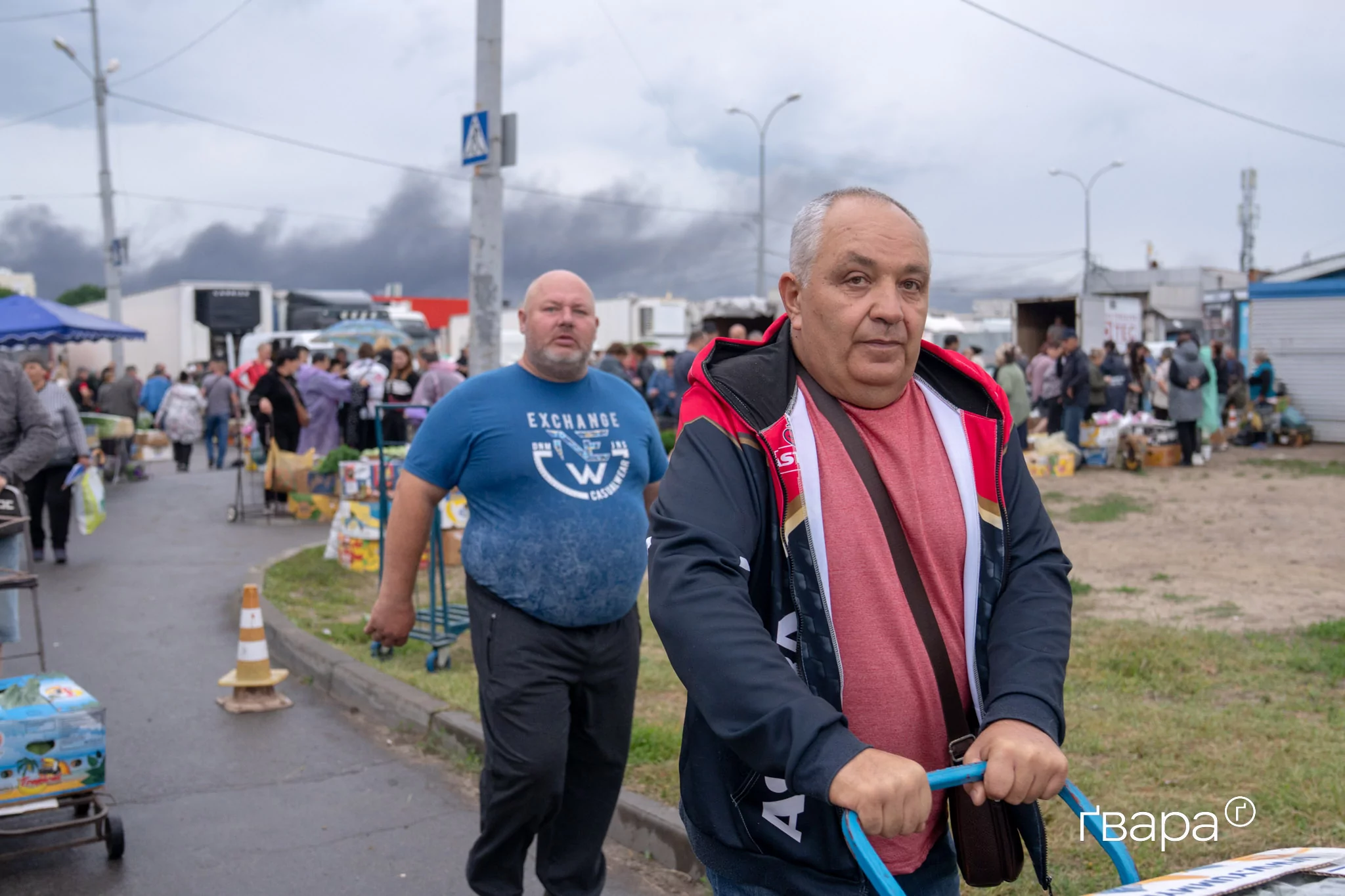 Люди купують продукти на ринку в Харкові на тлі диму від нічної російської атаки. Червень 2025 року
