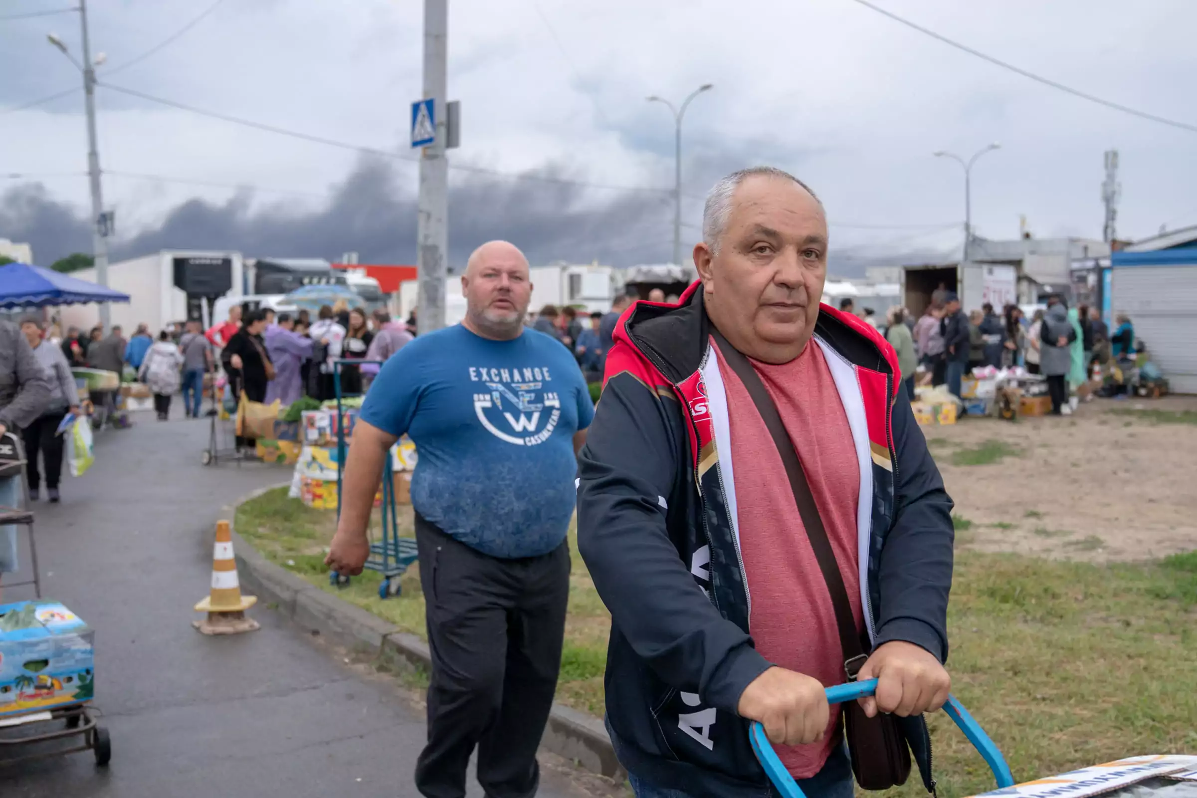 People are buying products at a farmer&rsquo;s market in Kharkiv with the smoke rising above the buildings after overnight Russian airstrikes. June 2025