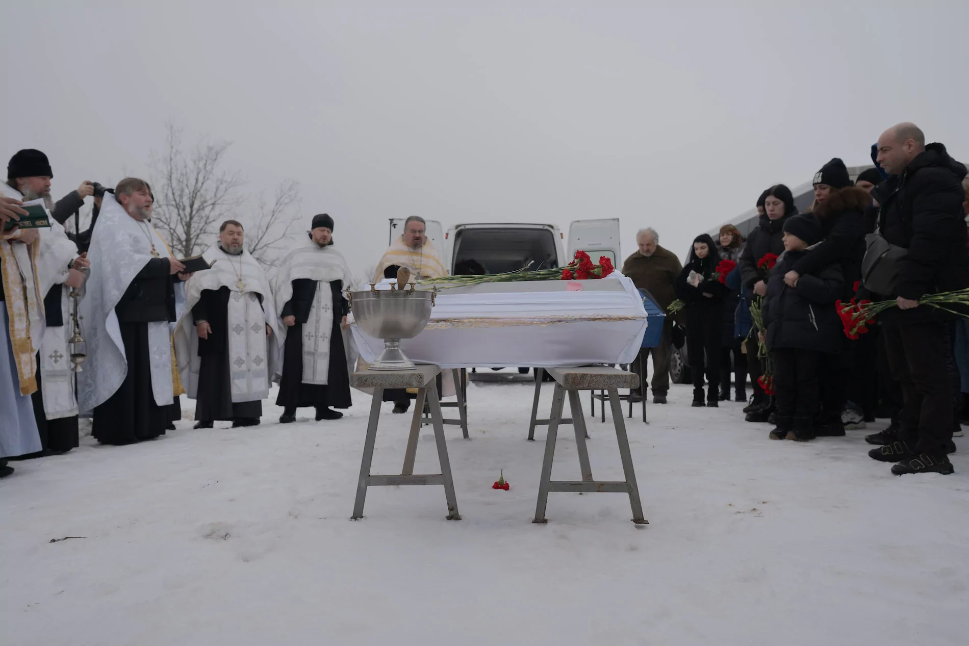 Vessel for holy water with aspergillum inside, behind him — a small white coffin. Priests to the left, people gathered for the funerals to the right.